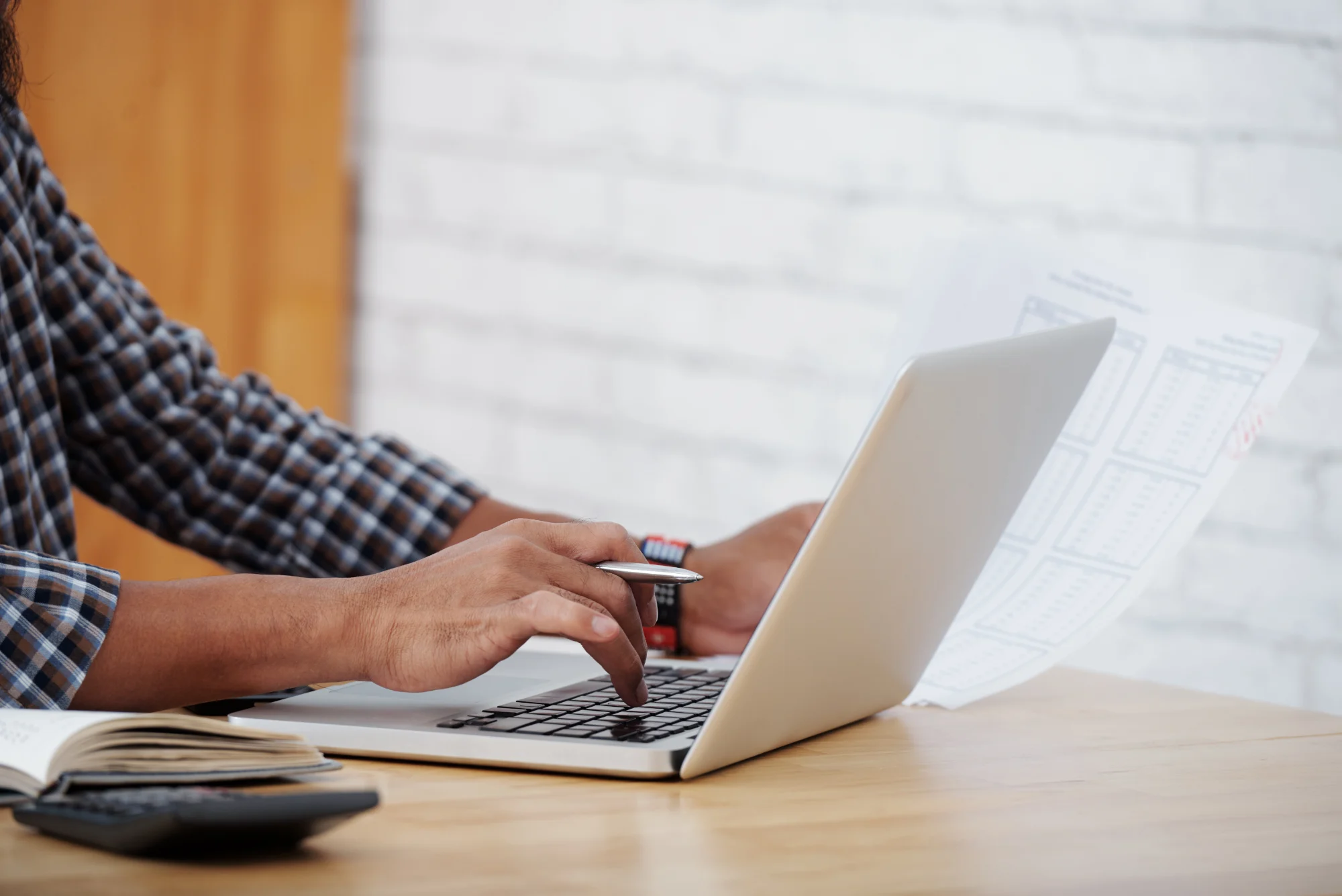 Hands typing on a laptop while holding a printed report, retyping figures by hand.