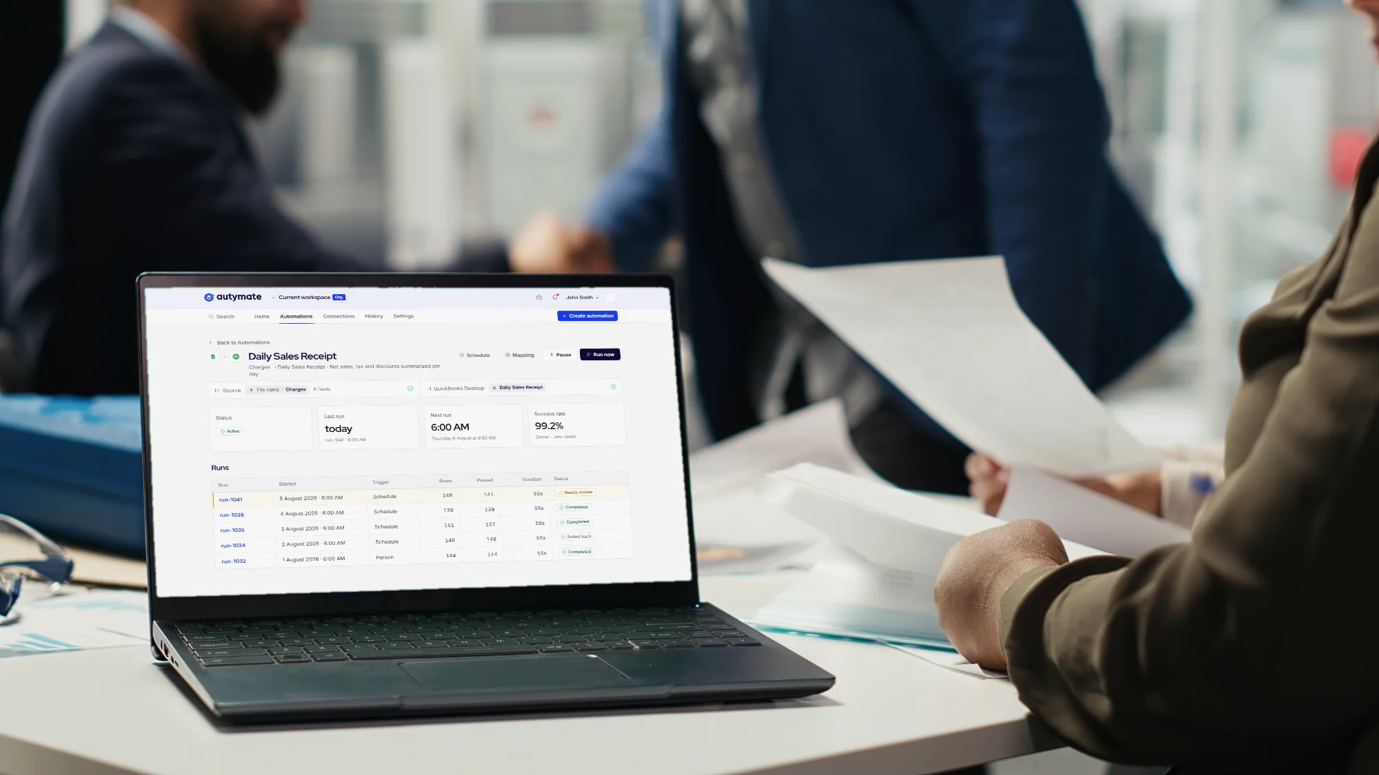 Two colleagues passing papers across a desk beside a laptop that shows an Autymate Daily Sales Receipt automation with QuickBooks Desktop as its destination.