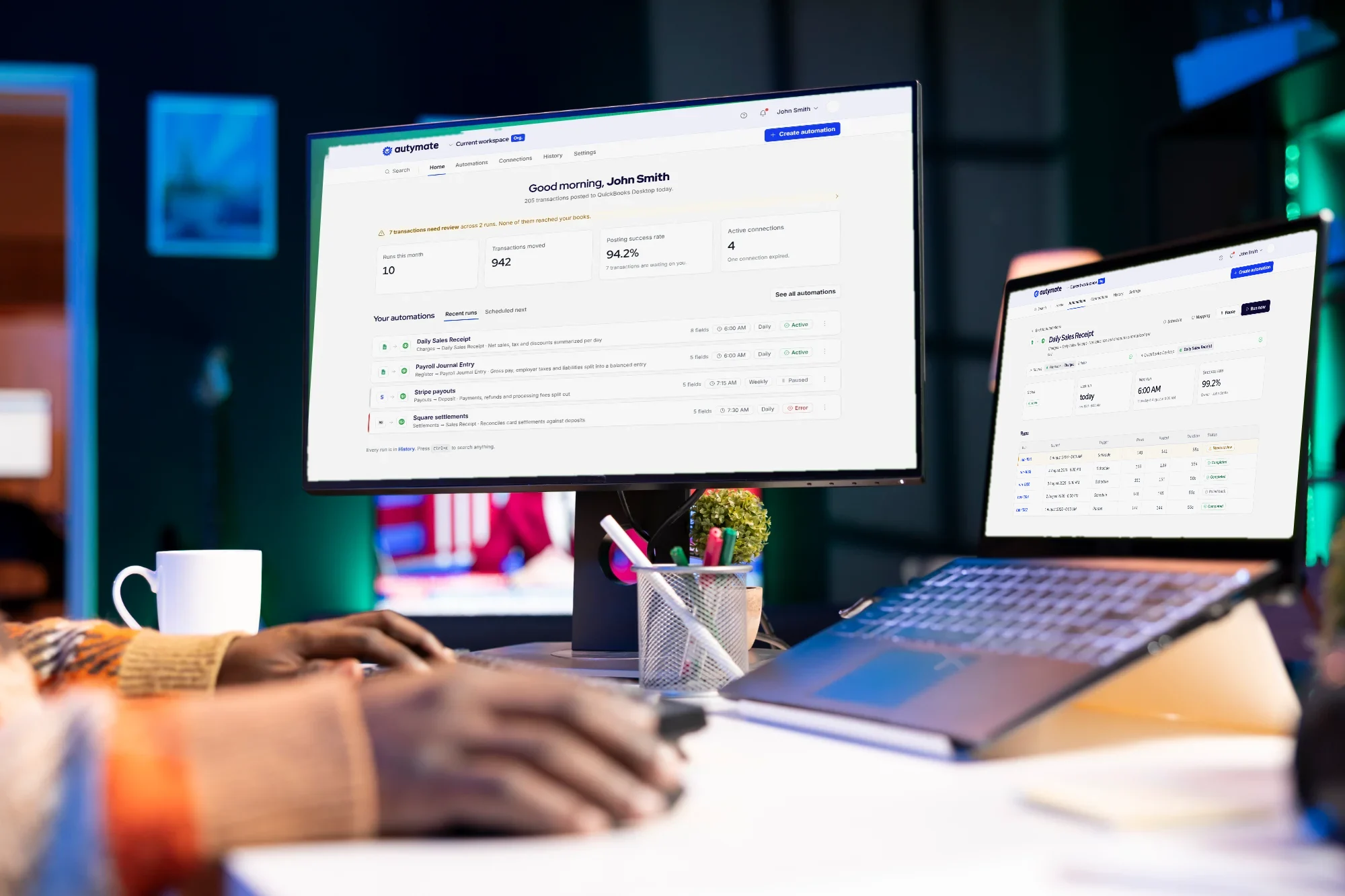 Hands at a keyboard in a dim office; the monitor shows the Autymate dashboard and the laptop shows the run history of a Daily Sales Receipt automation.
