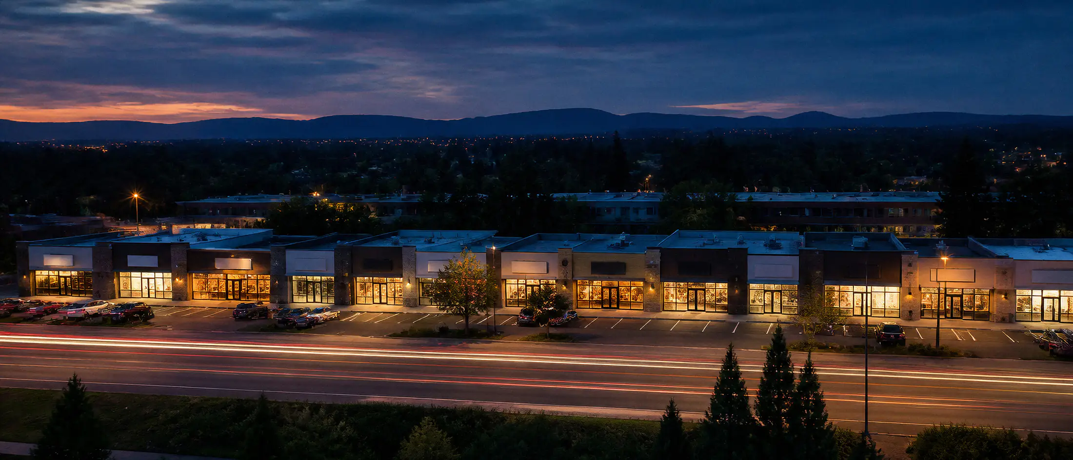 An elevated view at dusk of a retail strip of eight or nine similar single-storey units side by side, each one lit warm from inside, with a parking lot in front and traffic passing on the road below as streaks of light.