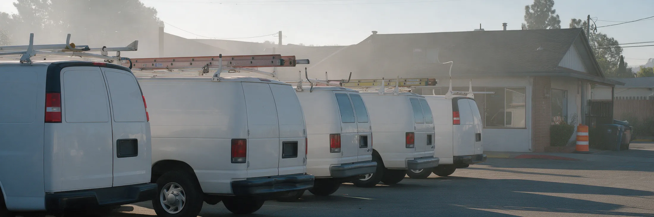 A row of unmarked white service vans with ladders on their roof racks, parked in a company lot at first light.