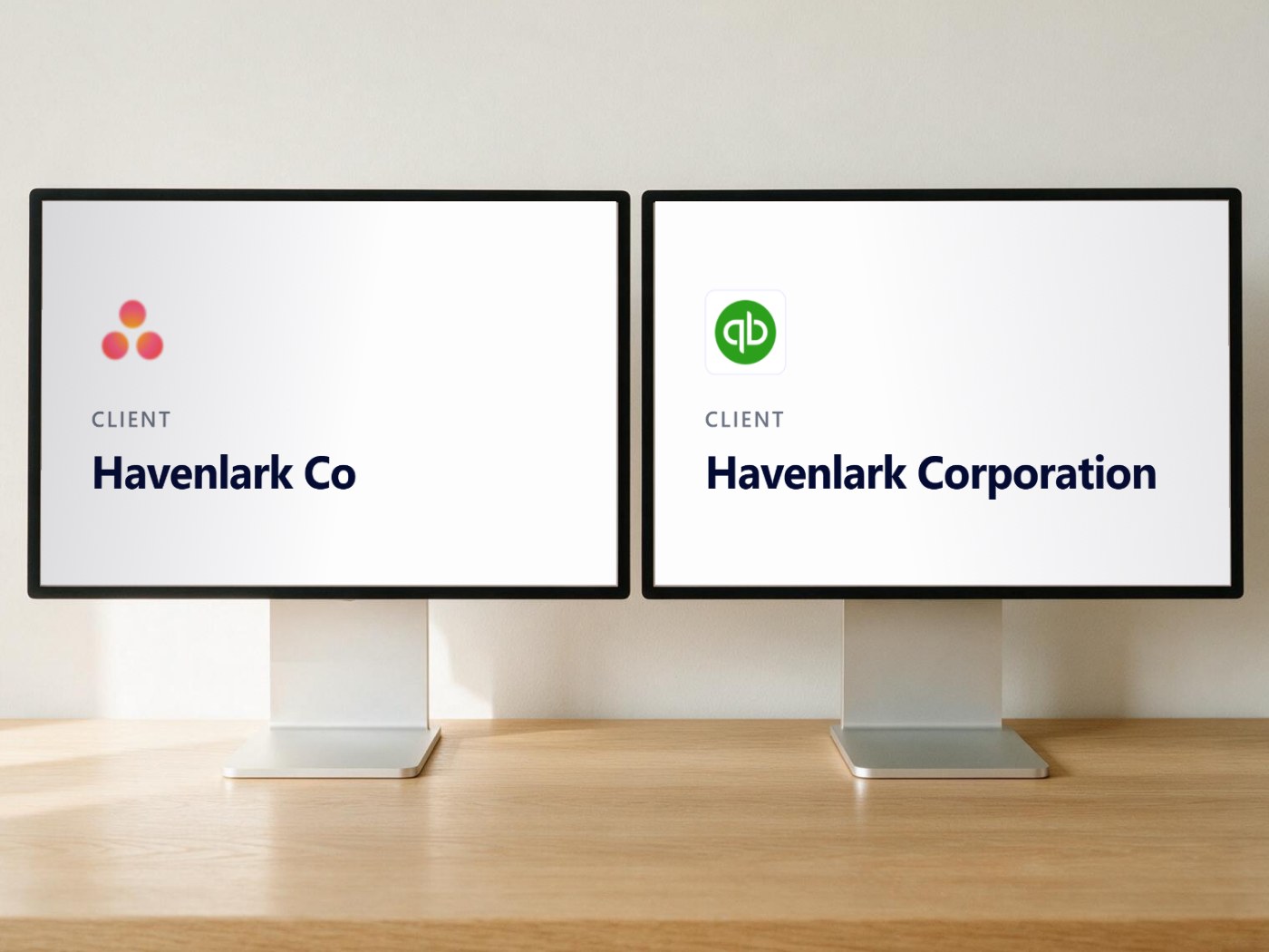 Two desktop monitors side by side on a wooden desk. The left screen shows the Asana logo above the client name Havenlark Co. The right screen shows the QuickBooks logo above the client name Havenlark Corporation. The same client, recorded under two different names.